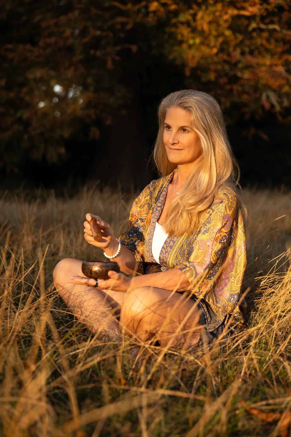 Carolyn Wade sitting in golden-hour grass, holding a singing bowl during a breathwork session.